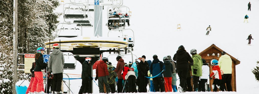 A group of people in winter clothes and on skis line up to wait for a ski lift to take them up the ski hill.