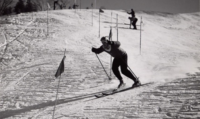 Black and white photo shows a skier turning a corner near a flag on a snowy ski hill.