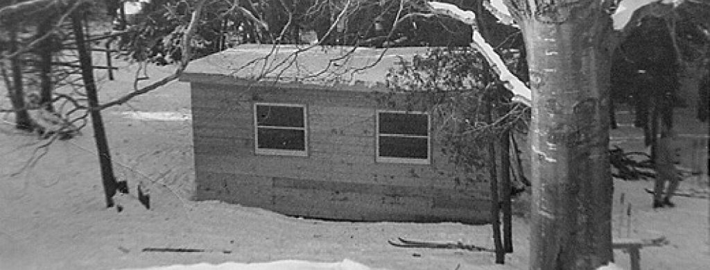 Black and white trail photo of a small cabin with two windows in the winter. The roof, trees and ground are covered in snow.