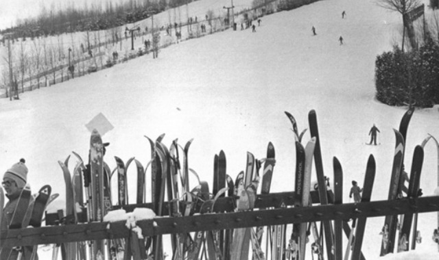 Black and white photo of a row of many skis available to use. In the background many people enjoy a ski hill.