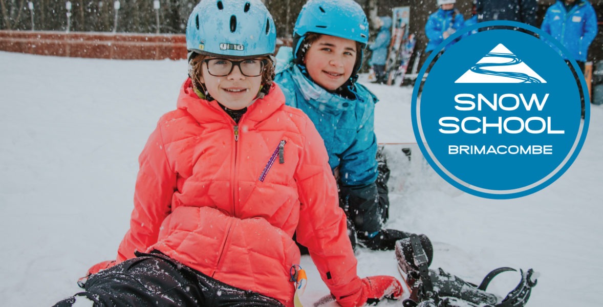 two young girls, wearing ski gear and helmets sit in the snow and smile