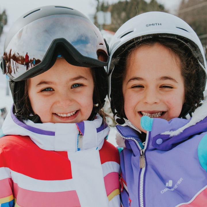 two young girls in ski helmets and jackets stand together and smile