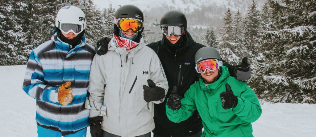 four men in ski gear stand together, smiling, and give thumbs up