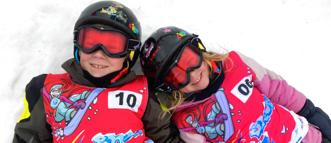 two girls lay together in the snow, wearing ski gear, and smiling