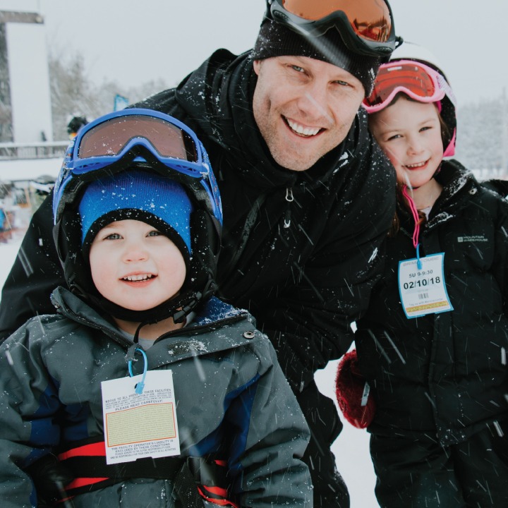 a father poses with his two kids, all are wearing skiing gear and are smiling