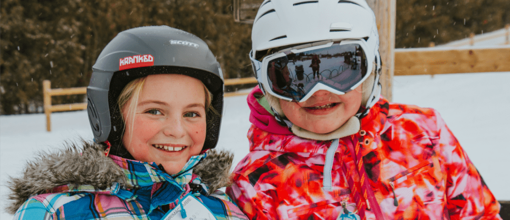 two young girls, wearing ski helmets and coats, smile for a photo